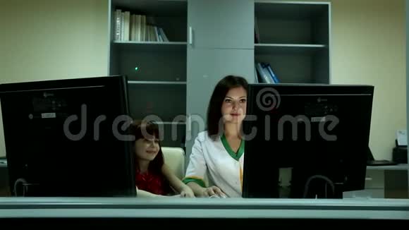 年轻漂亮的黑发女人医生看着两台电脑显示器后面的小可爱女孩微笑视频的预览图