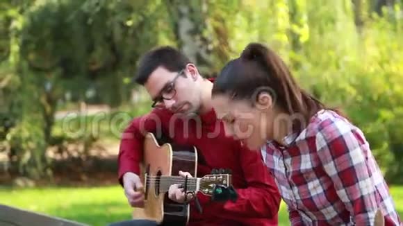 一对年轻夫妇坐在公园的长凳上男人弹吉他女人唱歌视频的预览图