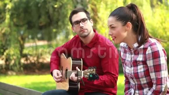 一对年轻夫妇坐在公园的长凳上男人弹吉他女人唱歌视频的预览图