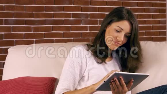微笑的女人在沙发上用平板电脑视频的预览图
