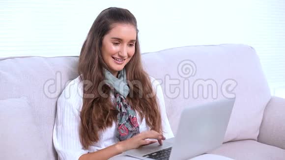 微笑的黑发女人用她的笔记本电脑在沙发上视频的预览图