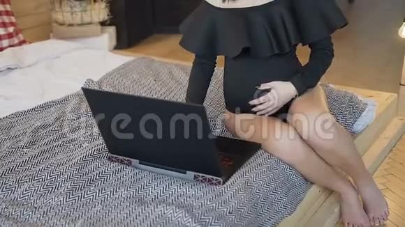一个美丽的怀孕的深褐色头发的女孩穿着黑色紧身西装坐在床上轻轻地摸着她的胃工作着视频的预览图