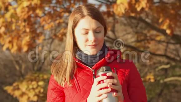 快乐的微笑女人享受大自然穿着红色外套带着热杯站在秋天公园的年轻女性视频的预览图