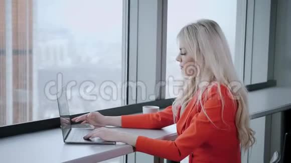 穿红珊瑚商务套装的女人年轻的金发女商人带着现代的灰色笔记本电脑在高层的办公室里视频的预览图