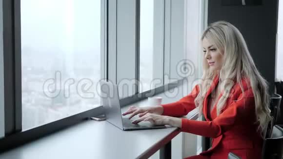 穿红珊瑚商务套装的女人年轻的金发女商人带着现代的灰色笔记本电脑在高层的办公室里视频的预览图