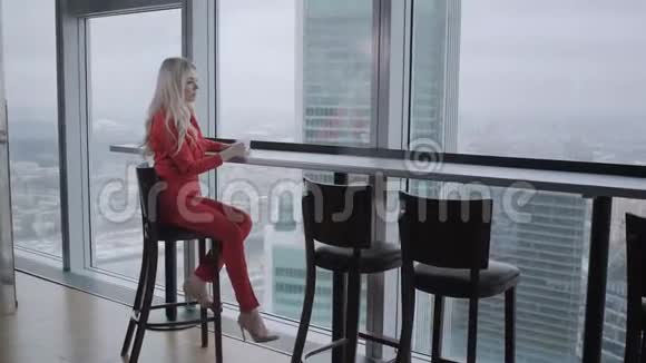 身着红珊瑚商务套装的女士在靠近窗户的高桌旁喝咖啡办公室里年轻的金发女商人视频的预览图