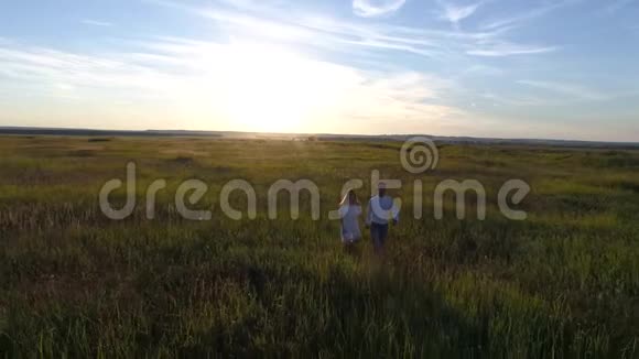 一对年轻夫妇穿过田野走向日落的空中摄影视频的预览图