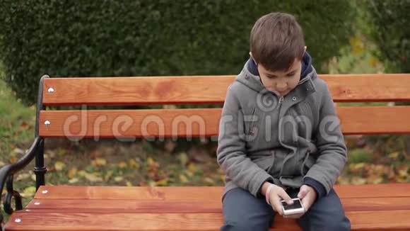 小男孩在公园里用电话他在秋天的天气里坐在长凳上背景黄树视频的预览图