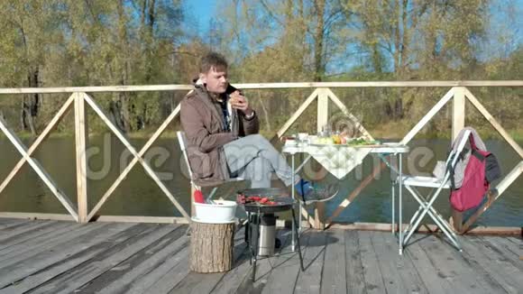 成熟的男人穿着温暖的衣服坐在桌子旁吃着汉堡靠近一只饥饿的狗在河岸上野餐视频的预览图