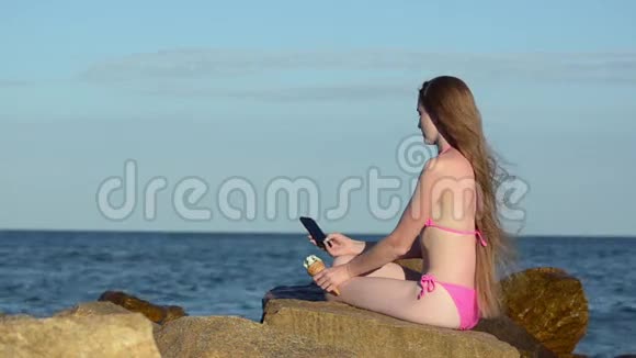 体贴的女孩穿着泳衣坐在海滩上的一块大石头上看着电话手里拿着冰淇淋风在你身上视频的预览图