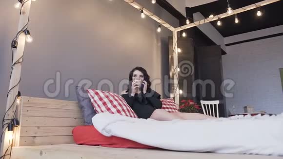 美丽的年轻孕妇坐在床上在一间时髦的卧室里用手机说话喝着热巧克力视频的预览图