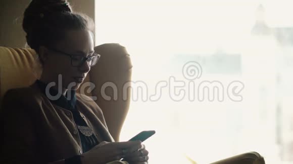 戴眼镜的年轻美女坐在窗户附近用智能手机上网视频的预览图