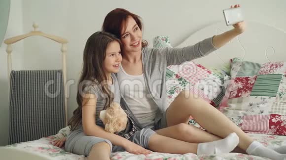 快乐的妈妈和小女孩用智能手机相机拍照坐在舒适的床上做鬼脸视频的预览图