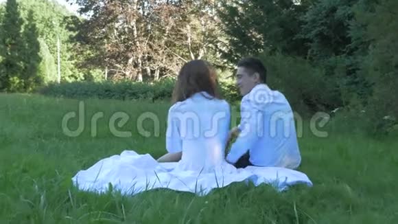 穿着白色衣服的浪漫夫妇拥抱并亲吻约会他们坐在一个美丽的公园里的绿草上视频的预览图