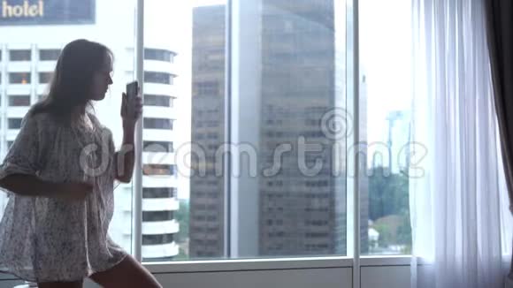 漂亮的黑发女人在全景窗户附近跳舞在公寓里用手机欣赏音乐和城市景色视频的预览图