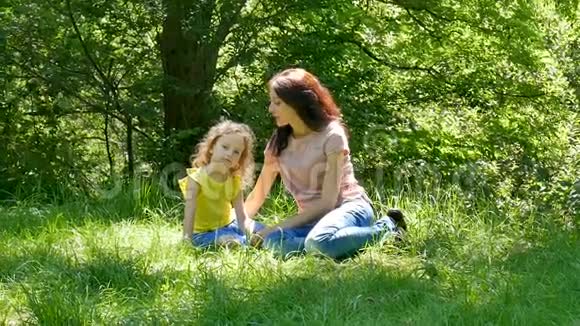 幸福的一家人坐在公园的绿草上小女儿在户外拥抱和亲吻妈妈视频的预览图