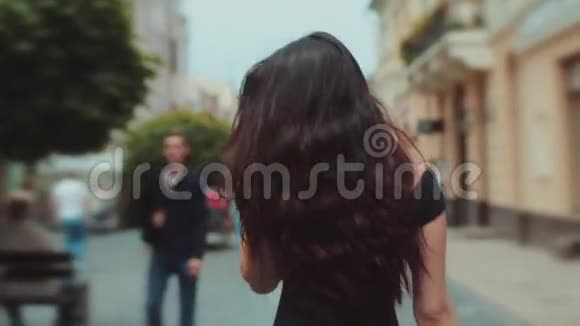 在城市的街道或小巷里走过的迷人的黑发卷发女人的后景转向相机和视频的预览图