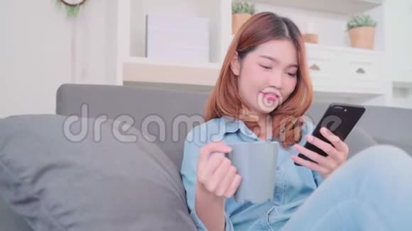 亚洲女性使用智能手机在家里客厅放松时一边躺在沙发上一边拿着一杯温暖的咖啡或茶视频的预览图