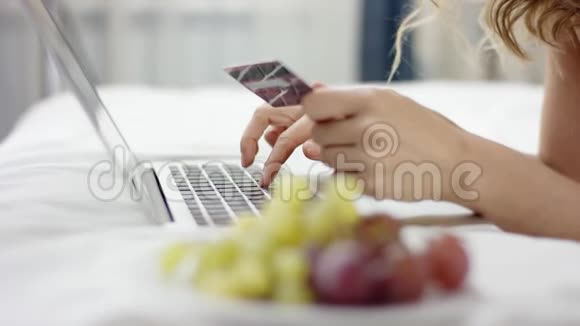 穿着睡衣的漂亮女人躺在床上用笔记本电脑和信用卡在网上购物视频的预览图