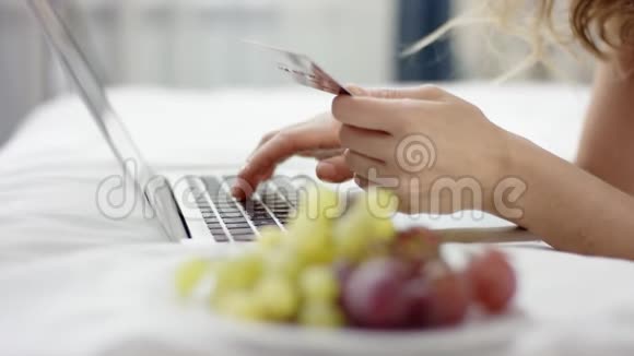 穿着睡衣的甜美女人躺在床上用笔记本电脑和信用卡在网上购物视频的预览图