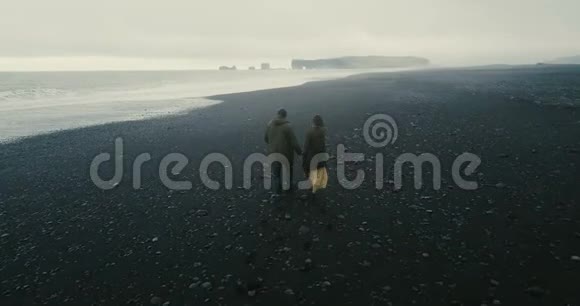 在冰岛黑色火山海滩散步的年轻夫妇的背面鸟瞰图海边的浪漫约会视频的预览图