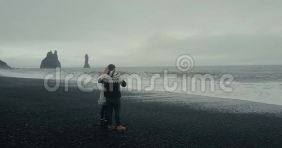 在冰岛年轻夫妇站在巨魔脚趾岩附近的黑色火山滩上看海浪视频的预览图
