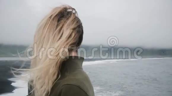 黑滩海岸上年轻的金发女人的后景在冰岛刮风的日子里看着巨怪的脚趾视频的预览图