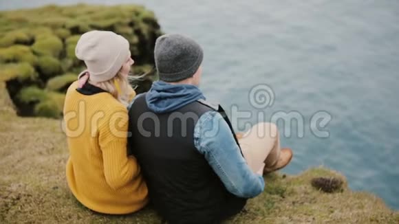 年轻漂亮的夫妇坐在悬崖边欣赏美丽的景色摄影师拍照视频的预览图