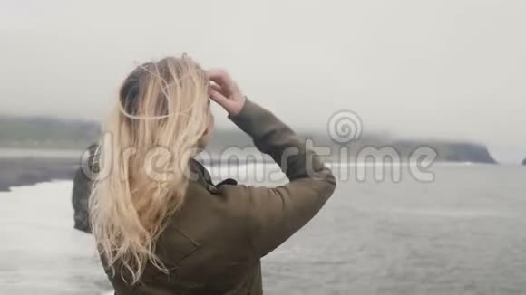 年轻的金发女人站在冰岛的黑色海滩上看着巨怪的脚趾岩石头发在风中飘扬视频的预览图