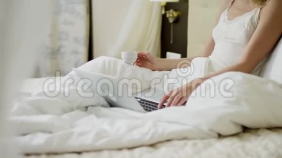 漂亮的女人一边在床上喝咖啡一边在笔记本上看新闻视频的预览图