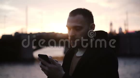 年轻留胡子的男人花时间在美丽的路堤上的肖像晚上在日落时使用智能手机视频的预览图