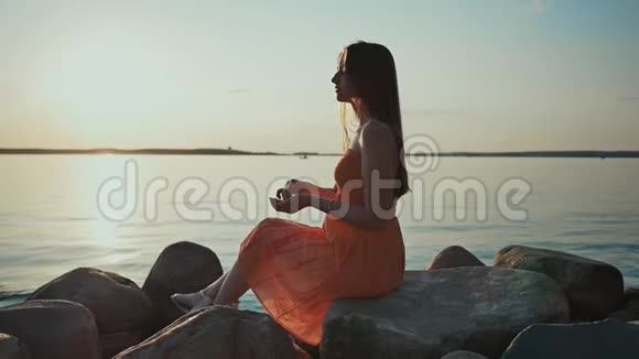日落时一个年轻的女孩坐在海滩上的岩石上扔石头在船帆的背景下视频的预览图