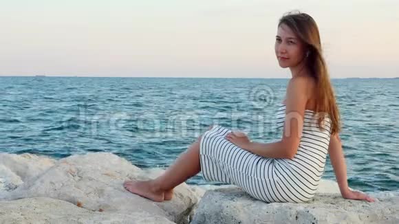 日落时分穿着白色连衣裙的年轻女孩坐在海边的石头上视频的预览图