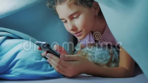 可爱的女孩和狗宠物的肖像在毛毯下与智能手机小女孩和狗宠物在网上玩游戏视频的预览图