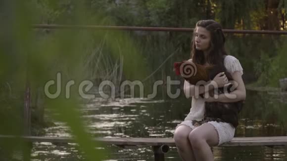 穿着白色衣服的女孩正坐在河上坐在木桥上拥抱公鸡视频的预览图