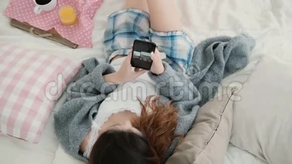年轻漂亮的女人躺在床上拿着手机女孩在智能手机上看旅行的照片视频的预览图