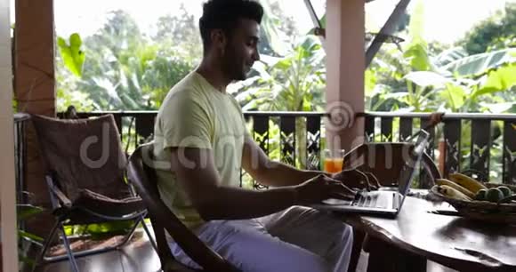年轻人在笔记本电脑上打字在露台上吃早餐在餐桌上工作时男人喝果汁视频的预览图