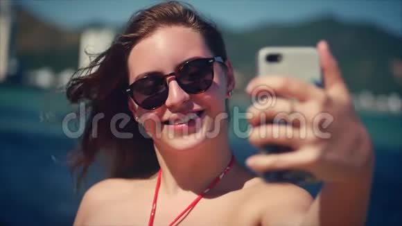 夏天年轻漂亮的黑发女人站在海滩上看着手机带着背景蓝色的自拍视频的预览图