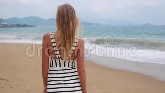 一个年轻的微笑女人穿着黑色和白色的裙子独自走在热带海滩上在背景上看着相机视频的预览图