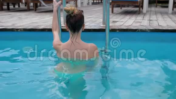 穿比基尼的迷人女人在蓝水背景下的梯子上从游泳池里爬出来年轻的女人视频的预览图