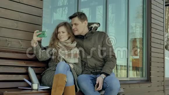 在一个坐在长凳上的春天公园里年轻的女人和男人在电话里自拍喝着热水瓶里的茶视频的预览图