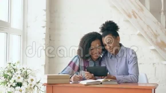 两个漂亮的卷发混血女孩坐在桌子旁一边学习和使用平板电脑一边玩得很开心视频的预览图