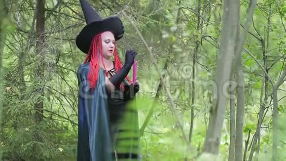 带着红色头发尖尖帽子和黑色斗篷的迷人的年轻女巫在森林里点燃蜡烛视频的预览图