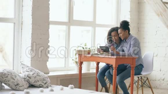 两个漂亮的卷发混血女孩坐在桌子旁一边学习和使用平板电脑一边玩得很开心视频的预览图