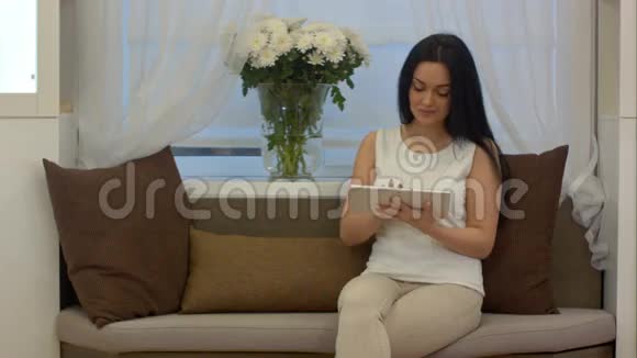 年轻漂亮的女人坐在沙发上在互联网上用触摸板搜索新闻视频的预览图