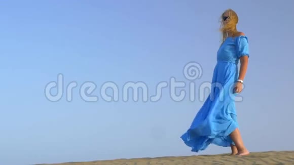 一个年轻的金发女子站在蓝色的天空上穿着蓝色的长裙在风中飘扬视频的预览图