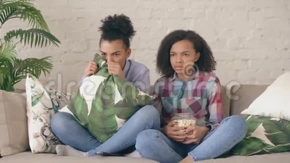 两个混血卷毛的女孩朋友坐在沙发上看电视上非常恐怖的电影在家吃爆米花视频的预览图