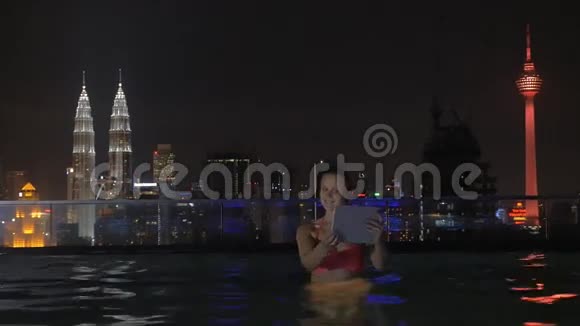 在摩天大楼屋顶上的游泳池里女人的视线通过平板电脑拍摄夜间城市景观吉隆坡视频的预览图