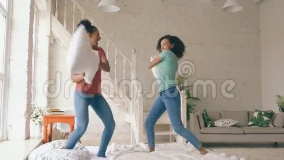 两个混合种族的年轻漂亮女孩在床上跳跃和打枕头在家玩得很开心视频的预览图