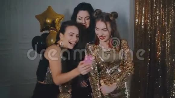 三个女孩参加派对金色背景气球女孩们在看智能手机上的照片笑着视频的预览图